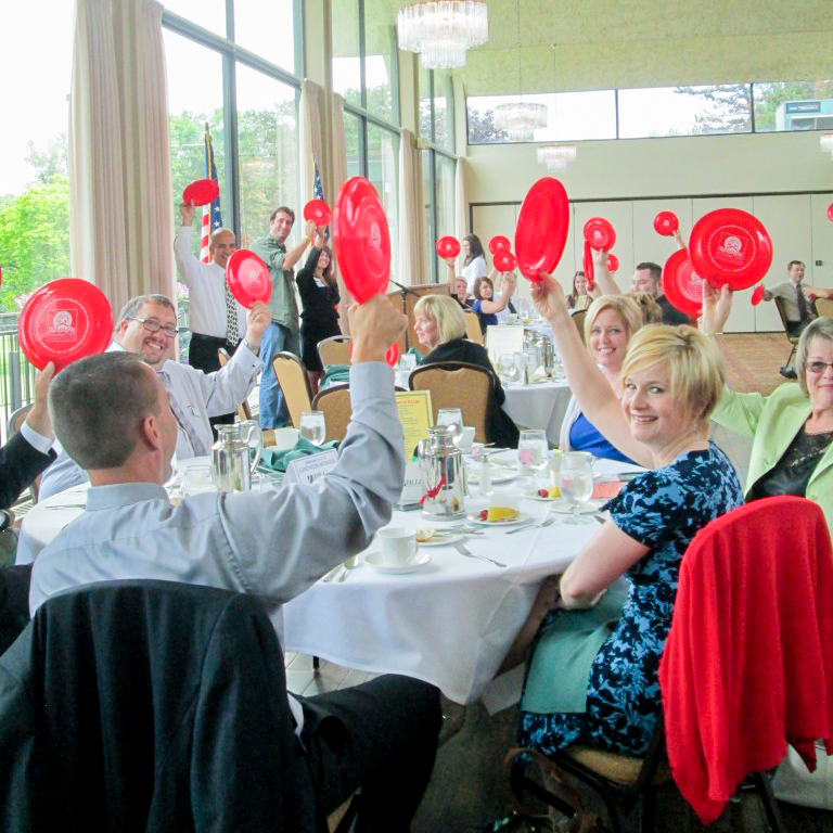 Many people seated at tables holding up a red frisbee